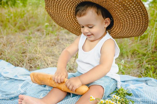 adorabile bambino ragazzo in cappello di paglia seduto sulla coperta al picnic e tenendo baguette
 - Foto, immagini