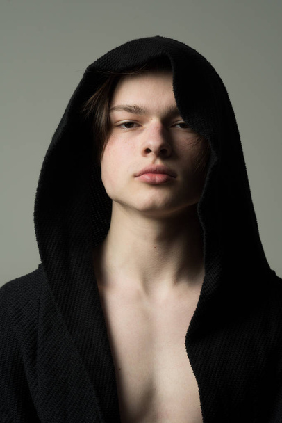 Portrait of young man wearing black hood, athlete or fighter before training, sports and health concept. Handsome guy in dark jacket over muscular torso isolated on gray background - Fotó, kép