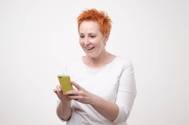 Close up portrait of businesswoman in white t-shirt and red short hair s looking at cell phone. She received interesting offer through sms - Photo, image