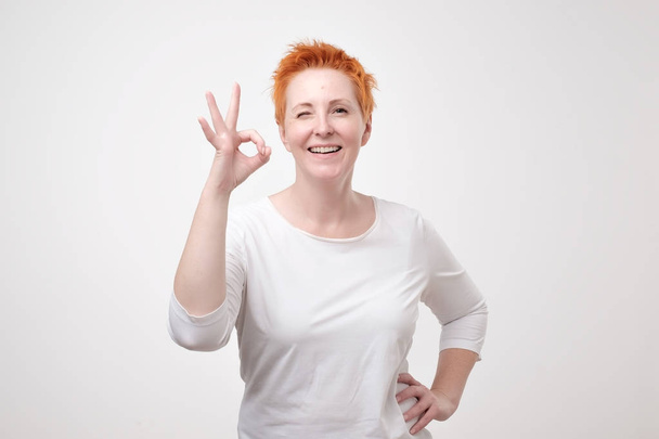 Усміхнена зріла леді в білій футболці жестикулює ідеальний знак. Вона задоволена результатом тренувань з фітнесу або косметичної процедури
 - Фото, зображення