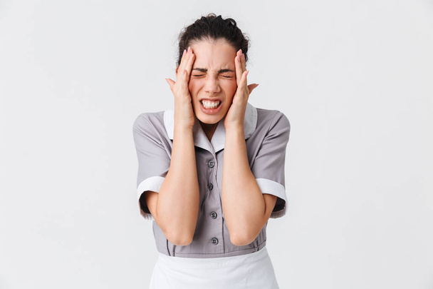 Portrait of an annoyed young housemaid dressed in uniform suffering from a strong migraine isolated over white background - Photo, image