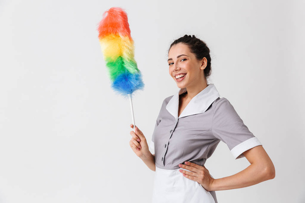 Portrait of a cheerful young housemaid dressed in uniform holding a duster isolated over white background - Foto, imagen