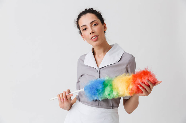 Portrait of a beautiful young housemaid dressed in uniform holding a duster isolated over white background - Foto, Imagem