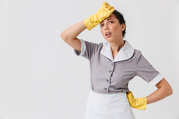 Portrait of a tired young housemaid dressed in uniform and rubber gloves wiping her forehead isolated over white background - Foto, Imagem