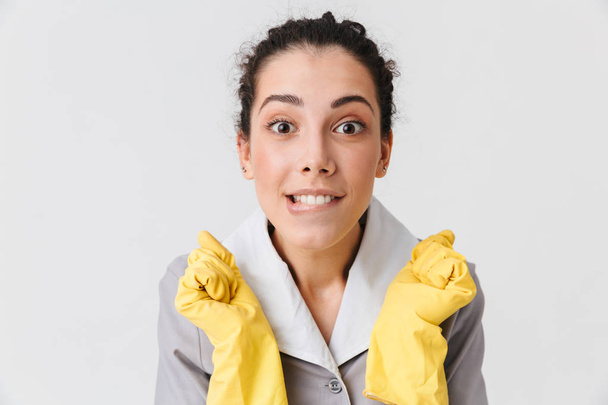 Portrait d'une jeune femme de ménage excitée vêtue d'uniforme et de gants en caoutchouc regardant la caméra isolée sur fond blanc
 - Photo, image