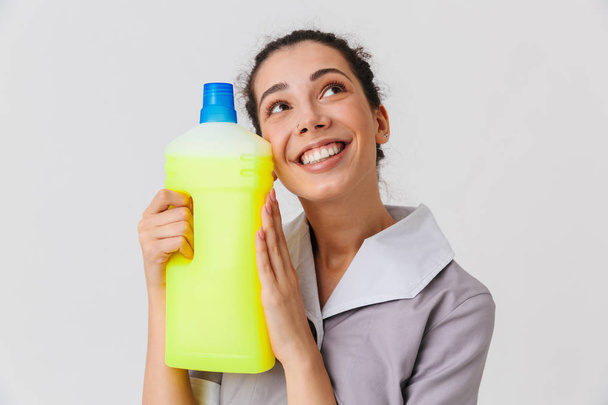 Portrait of a satisfied young housemaid dressed in uniform holding detergent isolated over white background - Fotó, kép