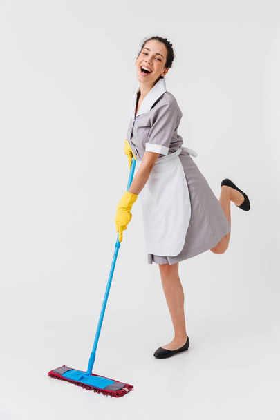 Full length portrait of a joyful young housemaid dressed in uniform using a mop isolated over white background - Фото, зображення