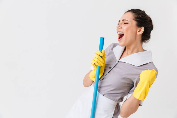 Portrait of an excited young housemaid dressed in uniform using a mop isolated over white background - Фото, изображение