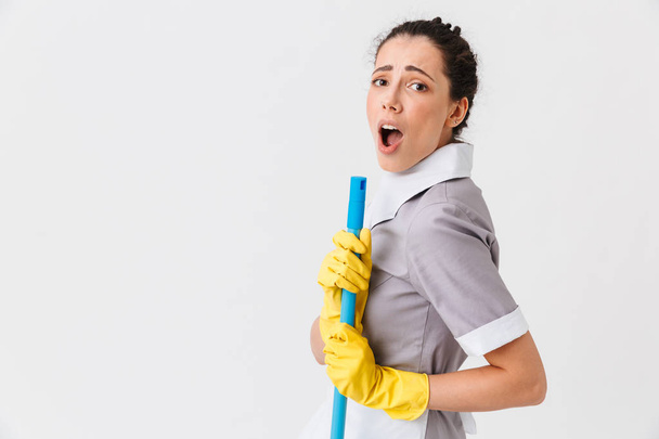 Portrait of a surprised young housemaid dressed in uniform holding a mop isolated over white background - Foto, Imagen