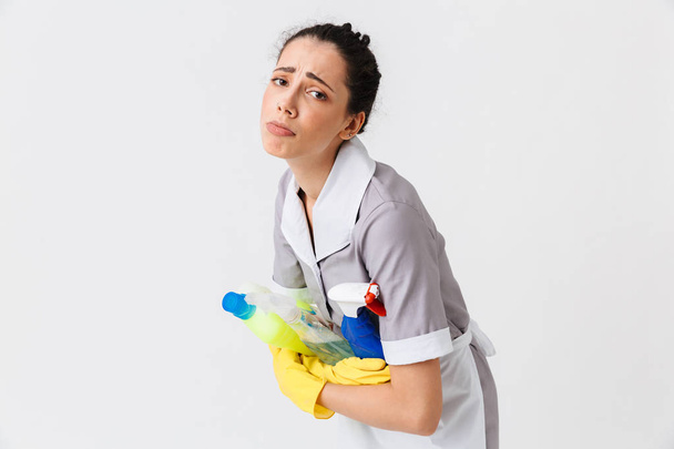 Portrait of a sad young housemaid dressed in uniform and rubber gloves holding detergents isolated over white background - Foto, Bild