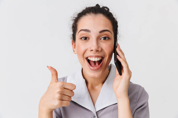 Portrait of a cheerful young housemaid dressed in uniform showing thumbs up while talking on mobile phone isolated over white background - Photo, Image