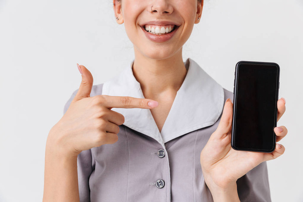 Cropped image of a cheerful young housemaid dressed in uniform pointing at blank screen mobile phone isolated over white background - Fotografie, Obrázek