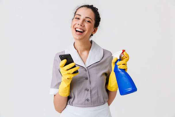 Portrait of a cheerful young housemaid dressed in uniform and rubber gloves using mobile phone while holding sprayer isolated over white background - Photo, image