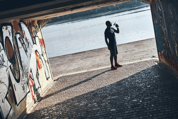 full length of young African man in sports clothing drinking water while standing at river  - 写真・画像
