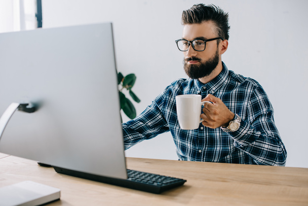 обрізаний знімок серйозного молодого сео з чашкою кави, що працює з комп'ютером в офісі
 - Фото, зображення