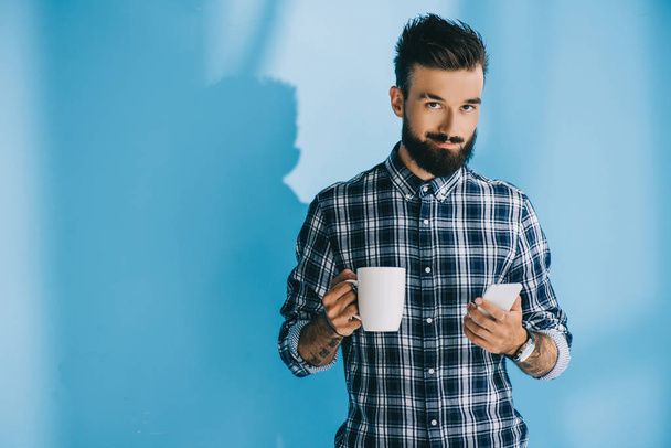handsome bearded man in checkered shirt holding smartphone and cup of coffee - Foto, Imagem