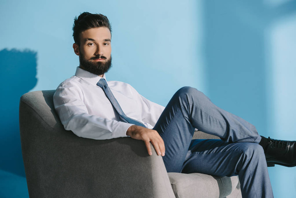handsome businessman in formal wear sitting in armchair, on blue - Foto, imagen