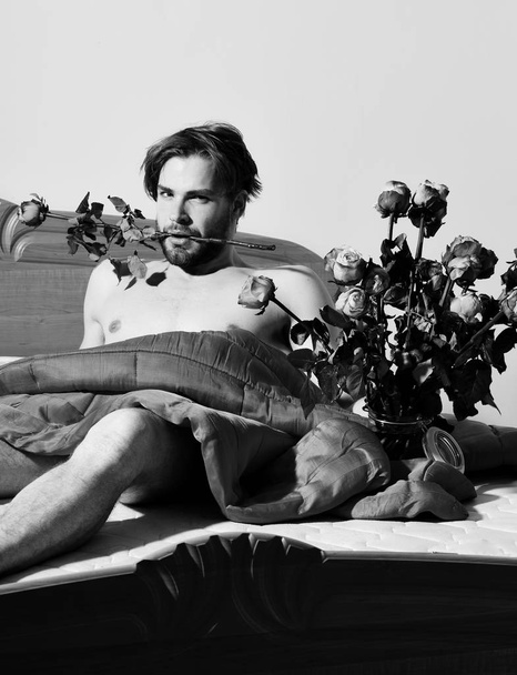 young handsome bearded sexy macho man with stylish beard lying naked on bed under red blanket holding bouquet of orange roses. - Photo, Image