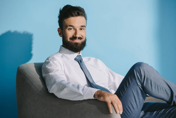bearded smiling businessman in formal wear sitting in armchair, on blue - Photo, Image