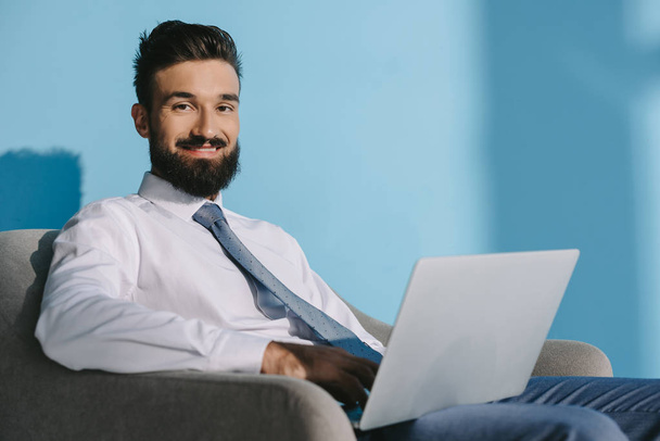 przystojny biznesmen uśmiechający się w stroje wizytowe za pomocą laptopa, na niebiesko - Zdjęcie, obraz