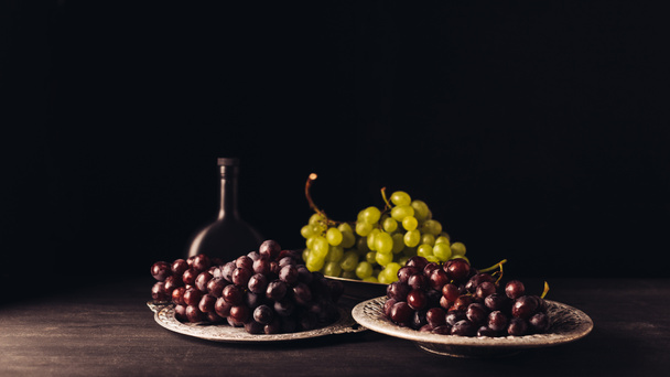 fresh ripe grapes on vintage plates and bottle of wine on wooden table on black   - Zdjęcie, obraz