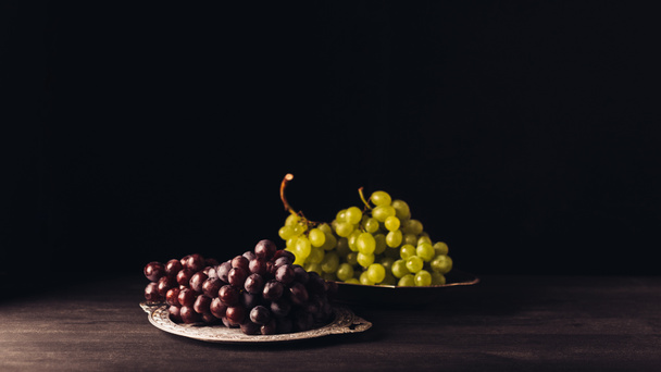 close-up view of fresh ripe red and white grapes on vintage plates on wooden table on black  - Foto, afbeelding