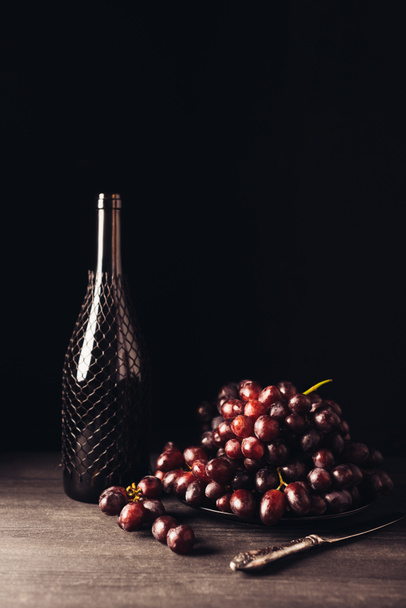 fresh ripe red grapes on plate, vintage knife and bottle of wine on wooden table on black - Foto, imagen