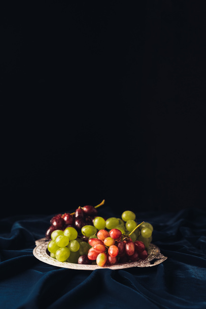 close-up view of various types of grapes on vintage plate on dark fabric on black background - Foto, afbeelding