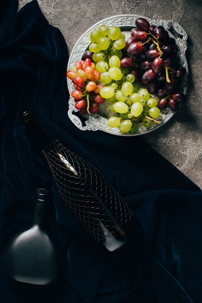 top view of various types of grapes on vintage plate and bottles of wine on dark fabric - Foto, Imagen