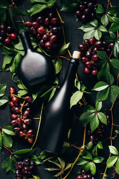 top view of fresh ripe grapes, green leaves and bottles of wine on dark wooden surface - Foto, afbeelding