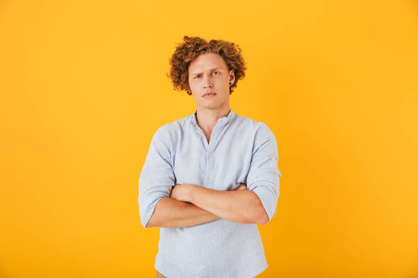 Photo of upset serious man 20s with curly hair standing with arms folded isolated over yellow background - Fotó, kép