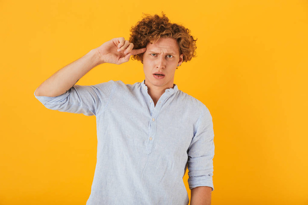 Photo of frustrated displeased man 20s with curly hair holding index finger at temple isolated over yellow background - Photo, Image