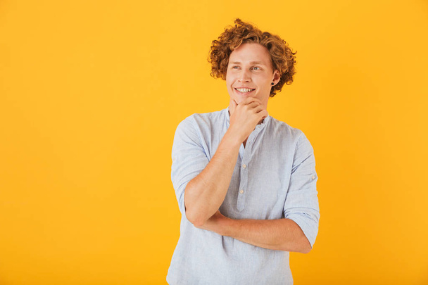 Portrait of happy caucasian man 20s touching chin and looking aside at copyspace with smile isolated over yellow background - Photo, image