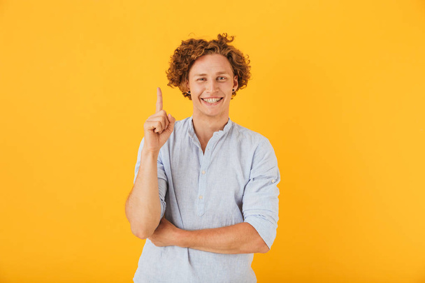 Portrait of joyful young man 20s smiling and pointing finger upward at copyspace isolated over yellow background - Foto, imagen