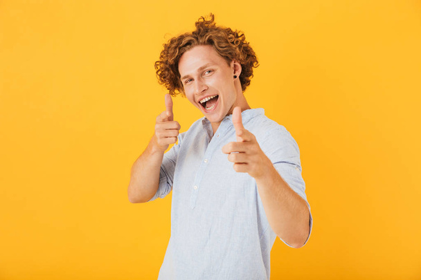 Photo of amusing handsome man 20s smiling and pointing fingers at you isolated over yellow background - Фото, изображение