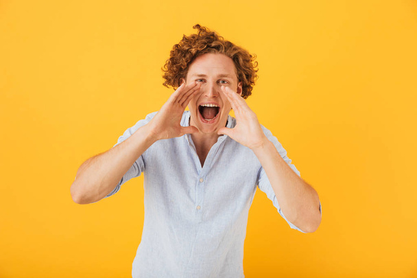 Photo of caucasian curly man 20s shouting or calling while holding hands at face isolated over yellow background - Photo, image