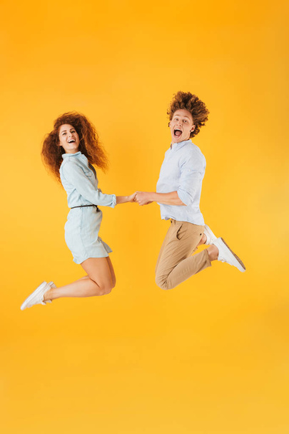 Full length portrait of excited couple man and woman 20s holding hands and jumping together isolated over yellow background - Photo, Image