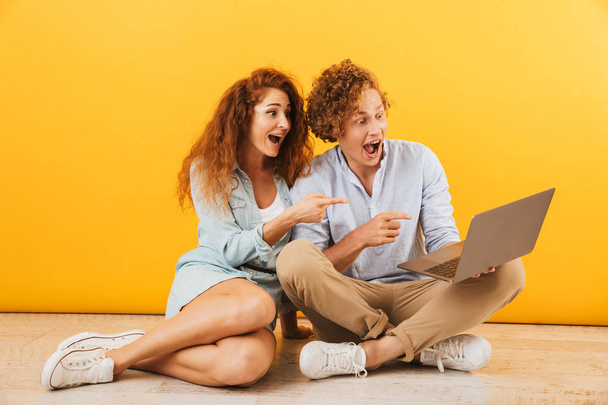 Photo of excited caucasian couple man and woman 20s sitting on floor and using silver laptop isolated over yellow background - Foto, imagen