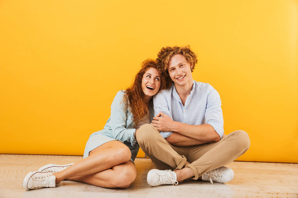Foto de optimista feliz pareja de hombres y mujeres de 20 años sonriendo y abrazándose mientras están sentados en el suelo juntos aislados sobre el fondo amarillo
 - Foto, Imagen