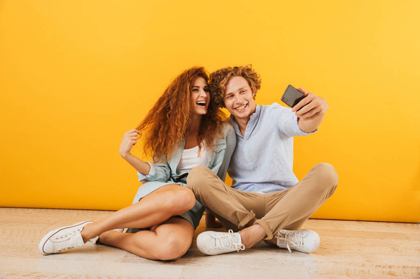 Photo of joyous boyfriend and girlfriend 20s sitting on floor together and taking selfie on smartphone isolated over yellow background - Fotó, kép