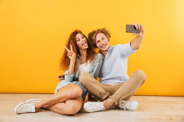 Photo of cheerful boyfriend and girlfriend 20s sitting on floor together and taking selfie on mobile phone isolated over yellow background - Photo, image