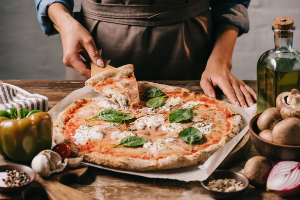partial view of woman in apron taking piece of pizza on cutting board on wooden tabletop - Foto, afbeelding