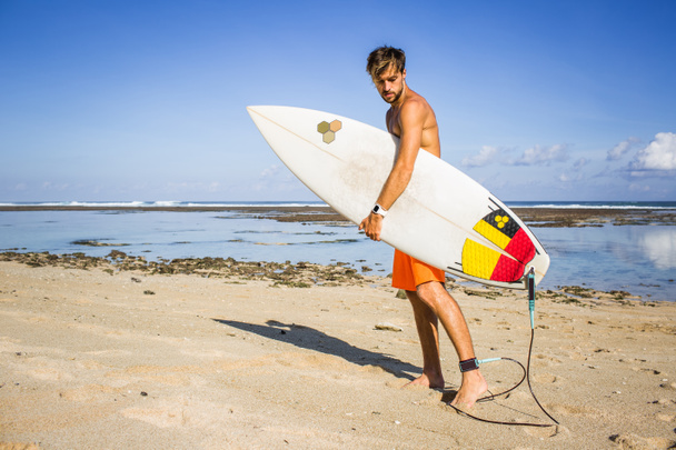 side view of young athletic sportsman with surfing board walking on sandy beach  - Photo, Image