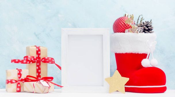 Bright christmas mock up with white horizontal photo frame: festive gift boxes, toys, and fir-cones in red santa's boot New year concept. Text space. Greeting card template - Fotografie, Obrázek