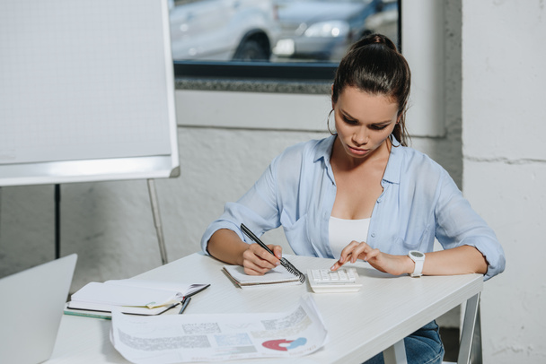 attractive businesswoman using calculator and writing something to notebook in office - Foto, Imagen