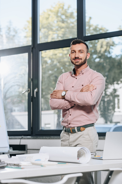smiling handsome businessman standing with crossed arms and looking at camera in office  - Zdjęcie, obraz