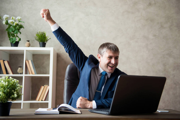 Happy excited young businessman sitting on workplace and celebrating success. - Φωτογραφία, εικόνα