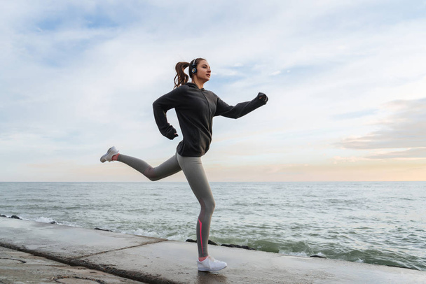 confident fast girl in gray sports clothes running around the sea in the evening, listening to music on headphones - 写真・画像