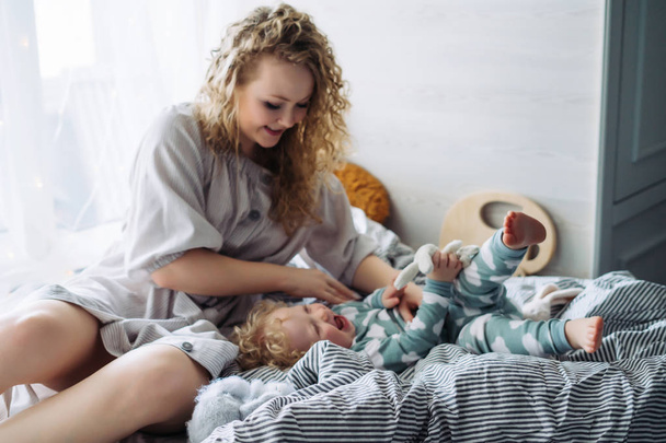 charming curly-haired young mother playing with her young son on the bed, the party is spending time - Zdjęcie, obraz