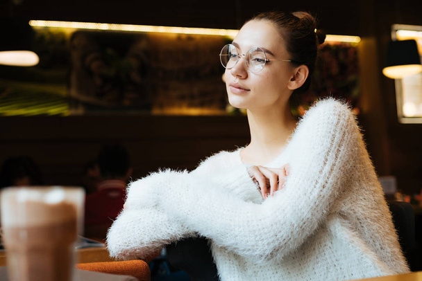 cute young girl student in a white sweater and glasses resting in a cafe, looking out the window - Photo, Image
